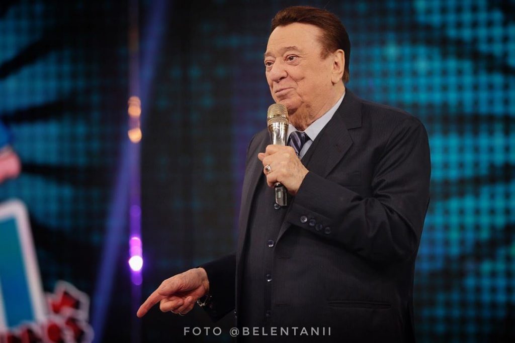 Foto colorida de Raul Gil, de terno, com um microfone na mão - Metrópoles