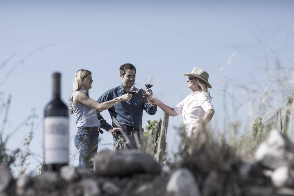 Três pessoas brincam e um vinho aparece em cima das pedras, a frente, em perspectiva