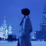 Na imagem colorida, o castelo de Moscou, na Russia está no fundo e uma pesso de lado e com roupas de frio ocupa o centro da imagem