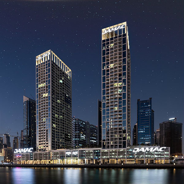 Prédios de luxo em Dubai, durante à noite. É possível ver a luz acessa dos apartamentos desses blocos. Eles estão localizados em frente a um rio.