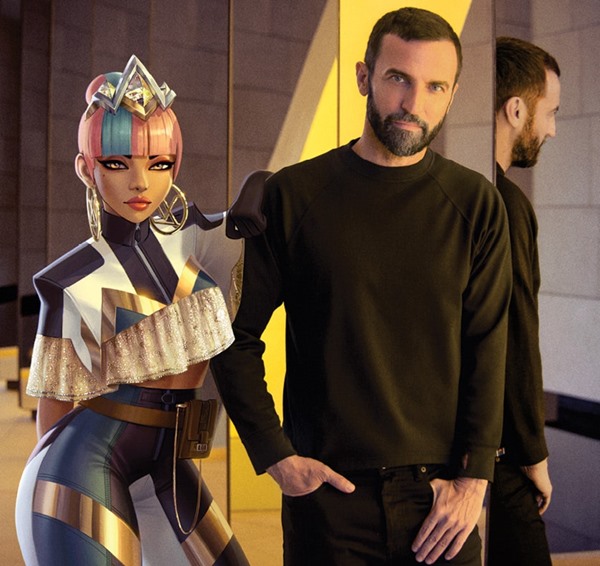 O estilista Nicolas Ghesquière posando ao lado de uma personagem virtual do videogame League of Legends Ele veste preto e a boneca animada possui o cabelo rosa e usa roupa dourada e azul
