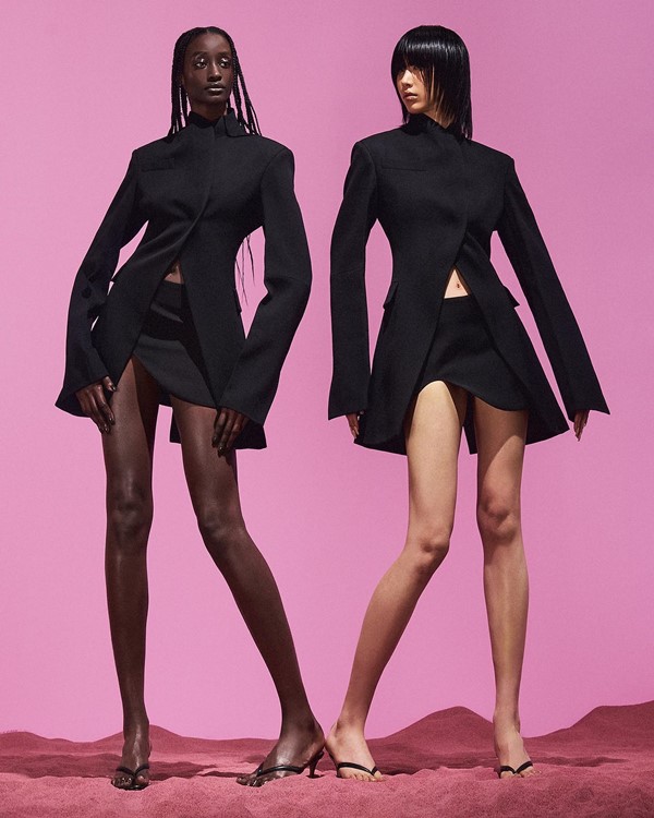 Duas mulheres posam em foto de campanha para a marca Maximilian Davis. A primeira é uma mulher negra e a segunda é asiárica. Ela usam saias e casacos pretos. O fundo da foto é rosa.