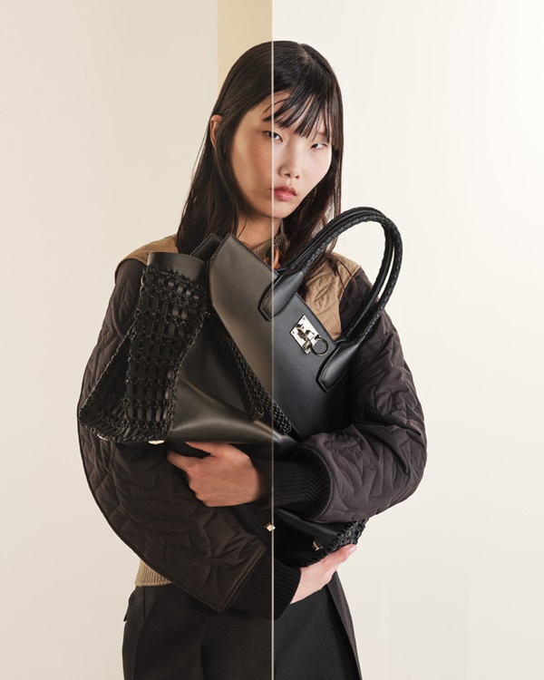 Mulher asiática com cabelo preto longo e de franja. Ela usa um casaco preto e segura uma bolsa de couro também na cor preta. As peças são da marca Salvatore Ferragamo.