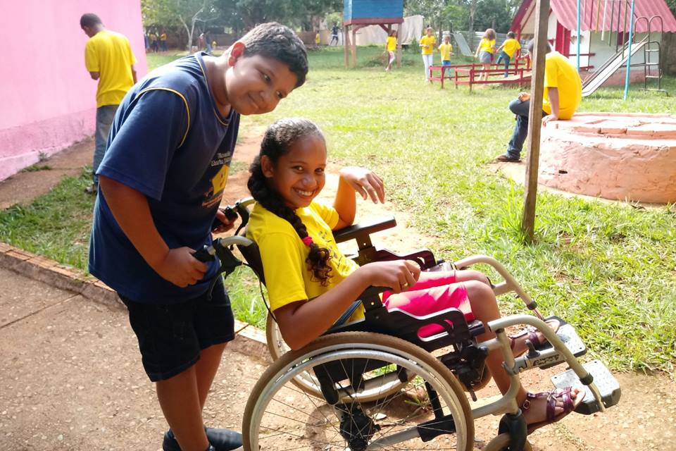 Foto colorida. Menino acompanha menina em uma cadeira de rodas