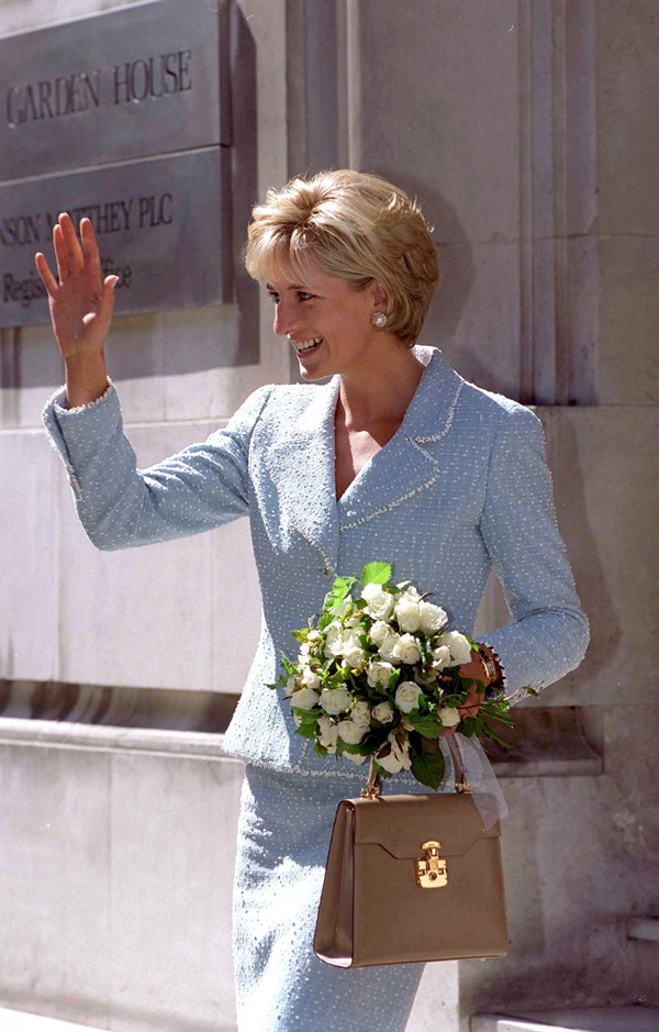 Princesa Diana usando conjunto de saia e blazer em azul claro. As peças são da marca Chanel. Ela segura uma bolsa bege e um buquê de rosas brancas em uma mão. Com a outra, acena para pessoas.