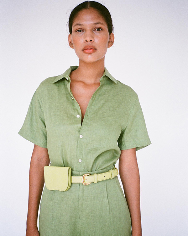 Mulher morena com cabelo liso amarrado para trás. Ela usa uma camisa de botão verde e um cinto com pochete em um tom de verde mais claro. As peças são da marca Pége.
