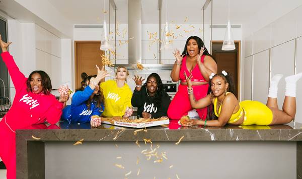 Cantora Lizzo e outras mulheres usando roupas esportivas em uma cozinha enquanto jogam batatas fritas para o alto
