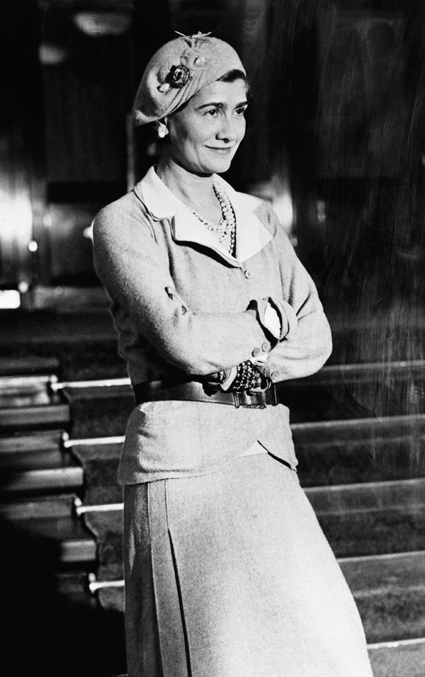 Fotografia da estilista Coco Chanel em 1926. Ela usa um conjunto de saia e blazer claro, chápeu e colares de pérola e de corrente, além de pulseiras. Ela está encostada em uma bancada com os braços cruzados.