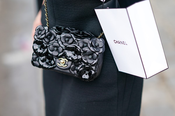 Bolsa preta da marca Chanel que imita a textura de várias flores Camélia. A pessoa segura uma sacola da mesma marca, a Chanel, como se tivesse saindo da loja - Metrópoles