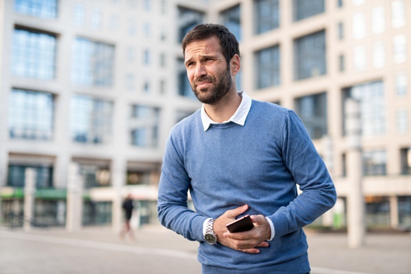 Homem em pé com uma das mãos na barriga e outra segurando um celular. Ele usa camiseta azul e tem cabelos e barba escuros - Metrópoles