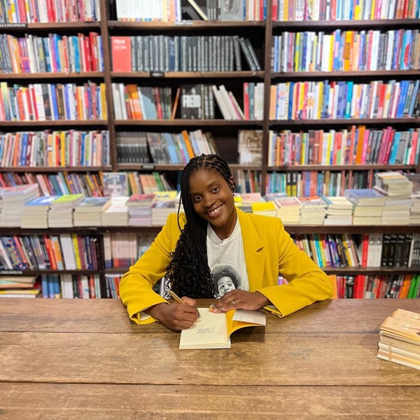 Mulher negra, com tranças, camiseta branca e um blazer amarelo sentada em uma mesa, em uma livraria. Ela está autografando o livro