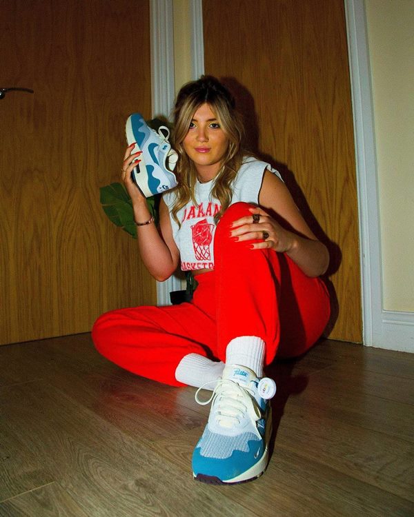Mulher branca e loura sentada no chão com calça vermelha, camisa branca e tênis com detalhes azuis da marca Nike