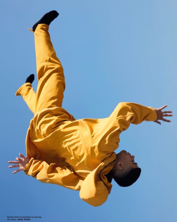Homem branco com cabelo preto fazendo um mortal no ar. Ele usa um conjunto de casaco e calça na cor amarela. A foto é uma campanha da marca Saul Nash.