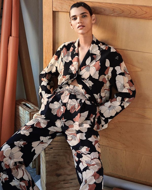 A modelo Kerolyn Soares, uma mulher de pele modera e cabelos curtos pretos, posando para foto em um fundo de madeira com rolos de tecidos azuis e marrons do lado. Ela veste um conjunto de blusa e calça floridos, com fundo preto, e flores brancas e rosas.