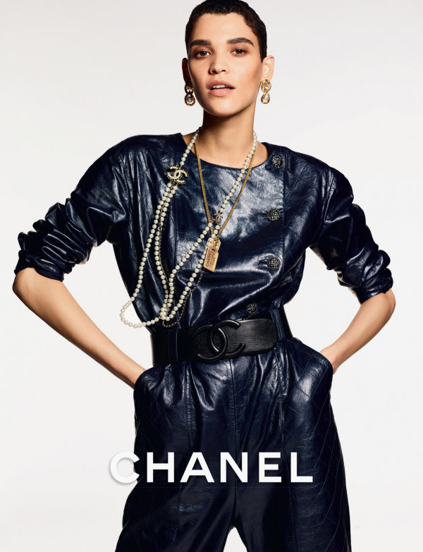 A modelo Kerolyn Soares, uma mulher morena com cabelo curto preto, em campanha para a marca de moda Chanel. Ela veste um macacão preto, de couro, com um cinto também preto com a logomarca da empresa. Usa correntes douradas e contas de pérola.