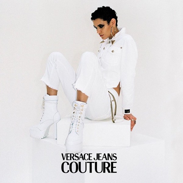 A modelo Kerolyn Soares, uma mulher morena com cabelo curto preto, em campanha para a marca de moda Versace. Ela veste uma jaqueta e uma calça jeans branco e uma bota, também branca, de salto.