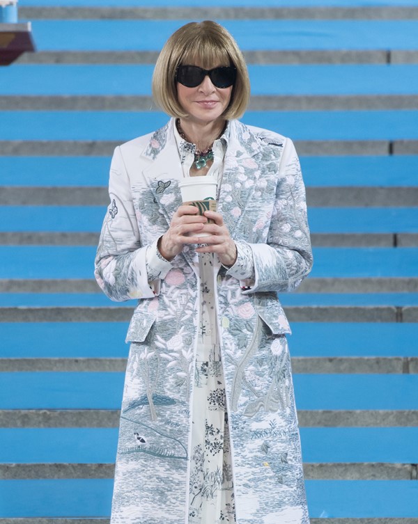 Anna Wintour, jornalista de moda e editora da revista Vogue, na escada do MET Gala em 2022. Ela é uma mulher idosa branca, com cabelo em corte chanel louro médio, e uma franja que cobre a testa. Veste um casaco branco com desenhos em azul, da marca Dior, e óculos escuros. Segura um copo de café da empresa Starbucks.