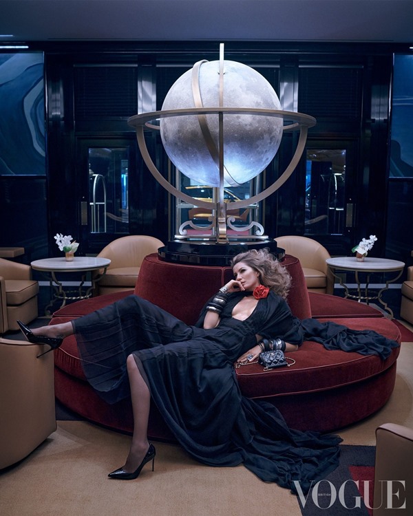 A modelo brasileira Gisele Bündchen, uma mulher branca com cabelo louro longo e ondulado, posando para a nova capa da revista Vogue da Inglaterra. Na foto em questão, ela está deitada em um sofá na cor vinho e usa um vestido preto, com salto alto também preto.
