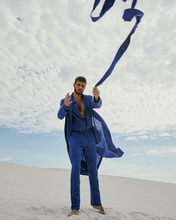 O ator Chay Suede, um homem branco com cabelo médio castanho, posando para foto em dunas de areia. Ele usa camisa de botão, calça e um kimono, todo em um tom de azul royal. A foto é de uma campanha publicitária da marca Handred.