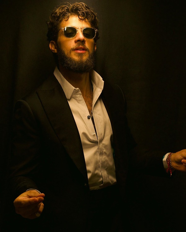 O ator Chay, um homem branco com cabelo e barba na cor castanha, posando para foto em um fundo escuro. Ele usa uma camisa branca, um terno preto e óculos escuros.