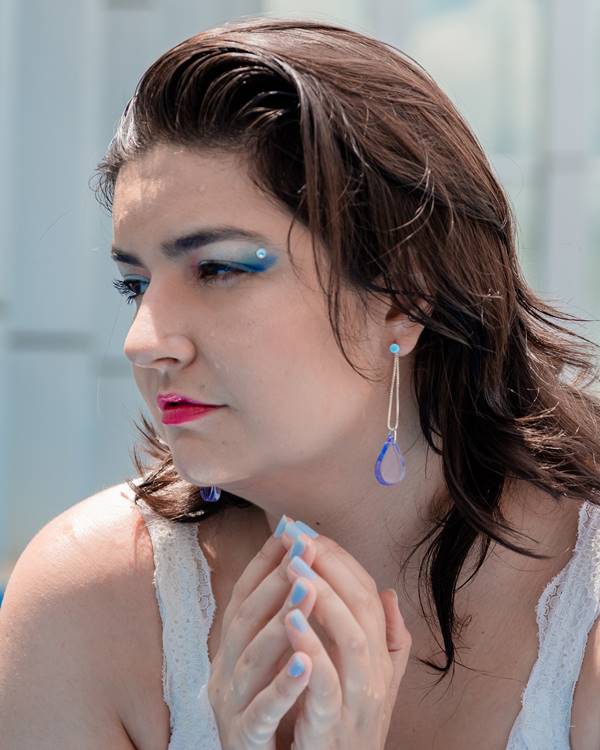 Mulher branca com maquiagem azul nos olhos mostrando brinco da marca Manualmente Joias