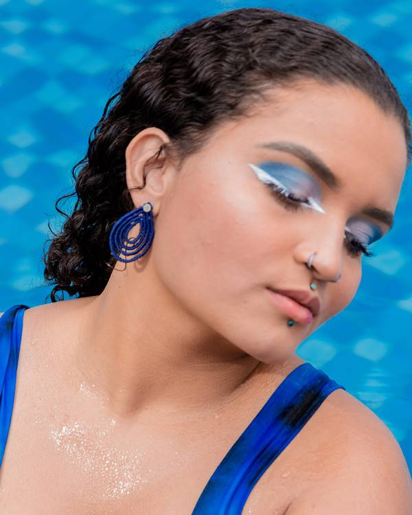 Mulher branca com maquiagem azul nos olhos mostrando brinco da marca Manualmente Joias