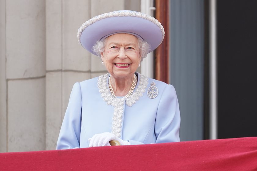 Rainha Elizabeth II está sorrindo para o público. Ela usa um conjunto roxo claro.