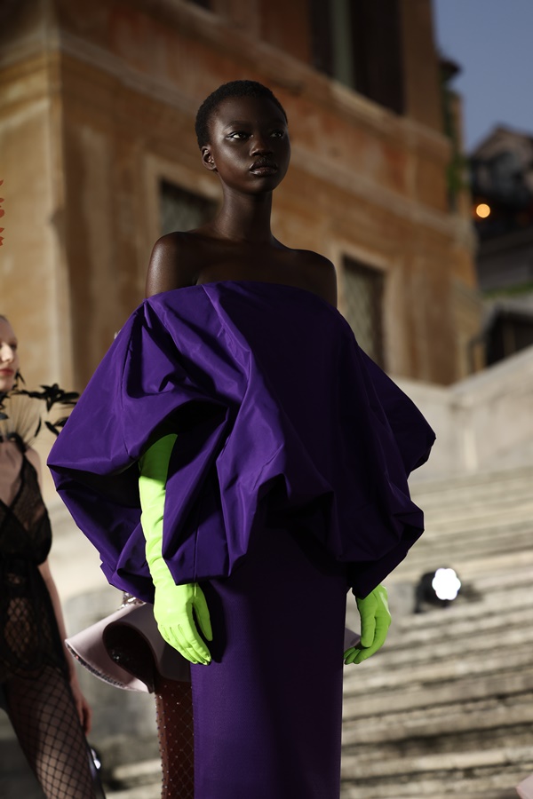 Modelo negra e jovem, com cabelo curto raspado, desfilando em escadas em uma praça de Roma, na Itália. É uma apresentação da marca Valentino e ela usa um vestido roxo com babados gigantes e luvas cumpridas em verde neon.