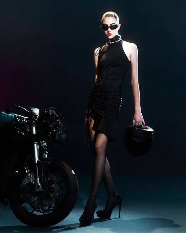 Modelo segura capacete com uma mão, ao lado de uma moto, usando vestido preto e curto de gola alta, em editorial da coleção Special, da Shop Ginger