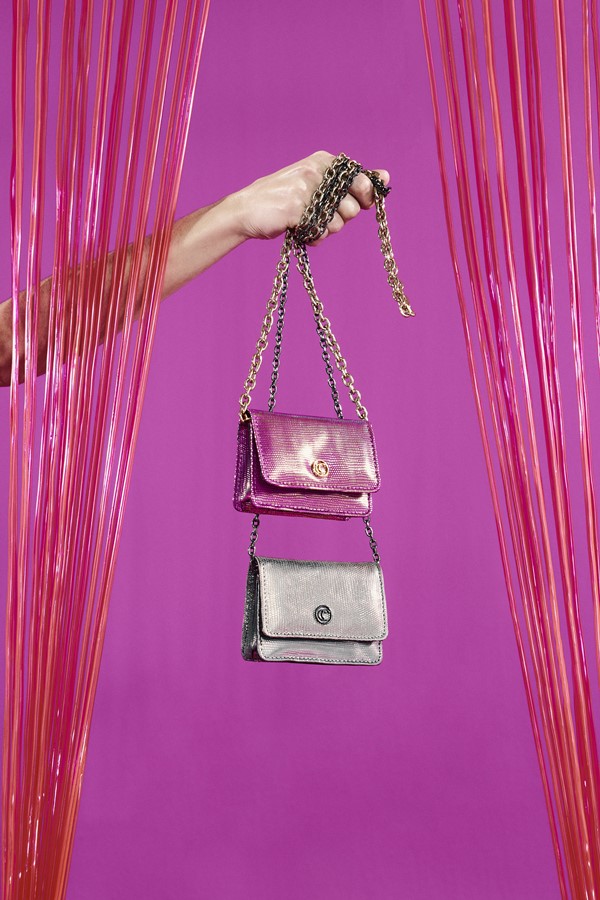 Campanha da nova coleção da marca Corello. Na foto, feita em um fundo rosa, é possível ver uma pessoa com a mão branca segurando duas bolsas da marca: uma rosa e outra prata.