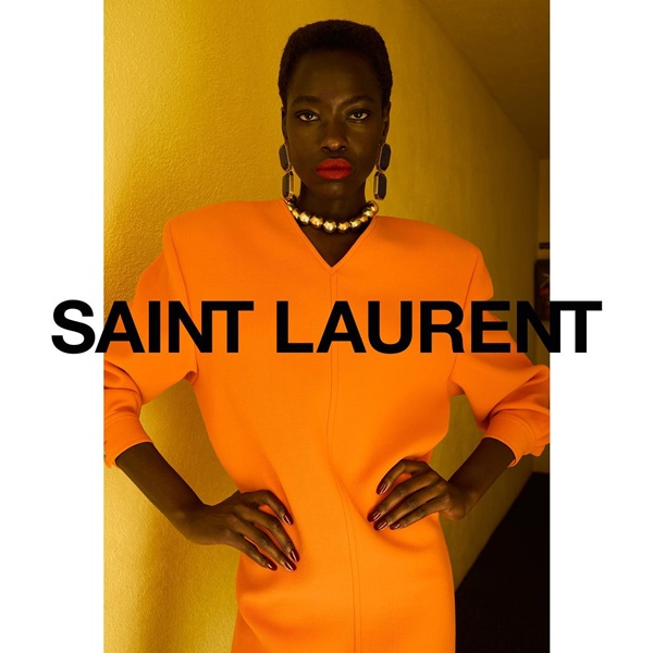 Campanha da marca de roupas de luxo Saint Laurent. Na foto, uma modelo, negra e de cabelo raspado, usa um vestido laranja de mangas cumpridas e com ombreira, um colar e um brinco grande.