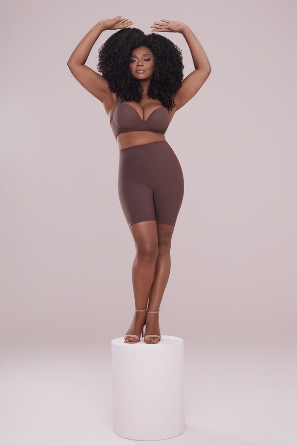 A cantora Paula Lima, uma mulher negra de meia idade com cabelos cacheados longos, posando para foto da campanha da marca de lingerie Plié. Ela usa um sutiã marrom e um shorts justo da mesma cor, além de sandálias de salto beges