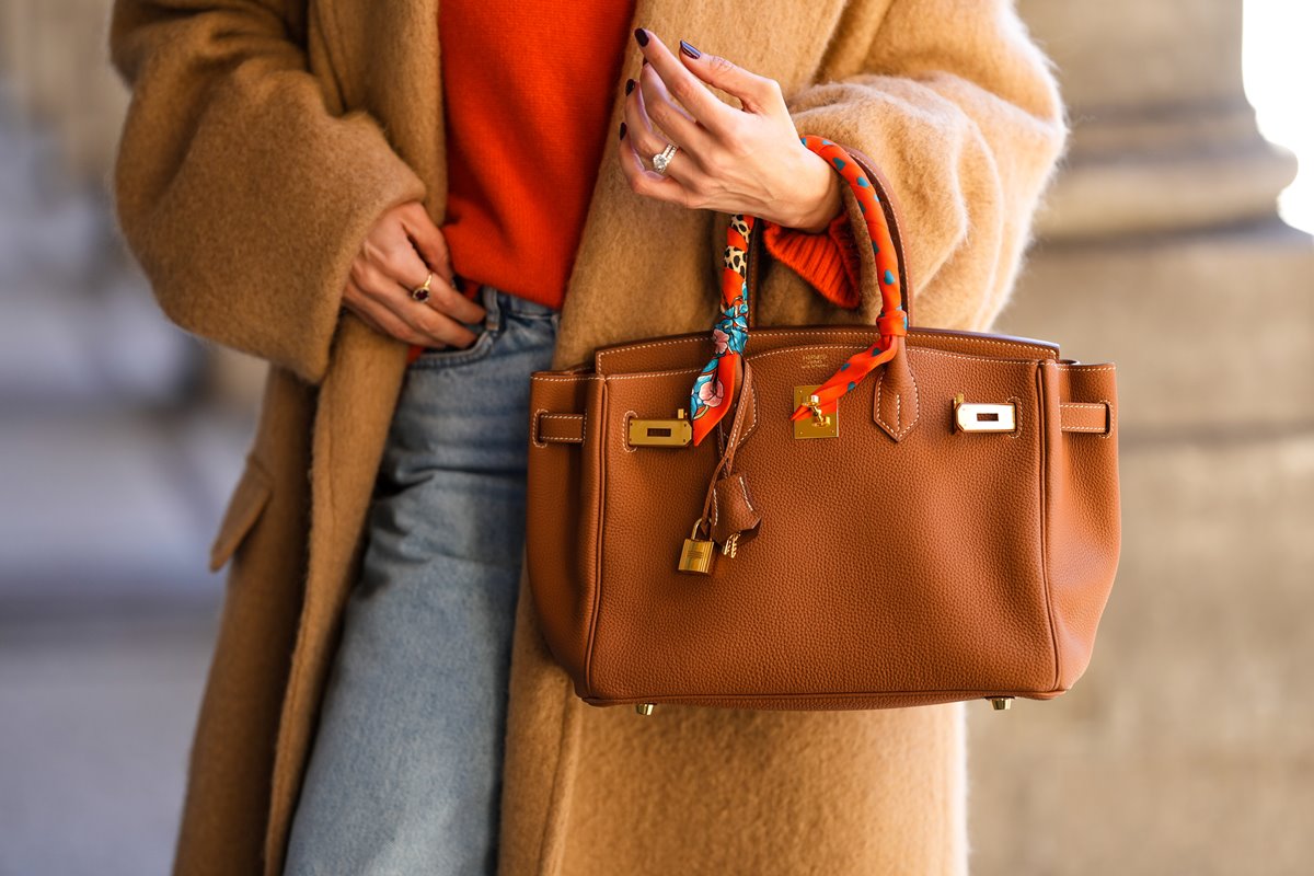 Mulher branca e jovem, segurando uma bolsa Birkin da marca Hermès em couro marrom. Ela usa um casaco de tricô laranja, uma calça jeans e um sobretudo de pelo bege.