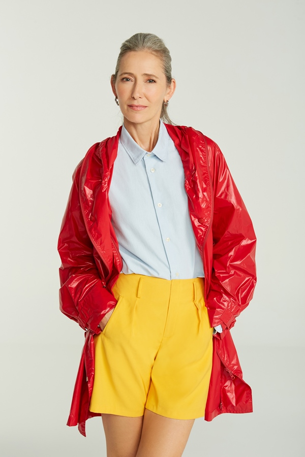 Campanha de divulgação da Simples, submarca da Reserva, onde modelos posam com as roupas em um fundo branco. Na foto, uma mulher idosa, de pele branca e cabelo longo grisalho, com uma camisa de botão, um shorts amarelo e um casaco vermelho.
