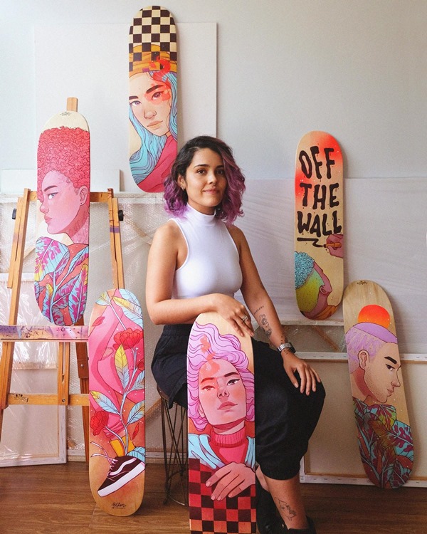 Mulher branca e jovem, de cabelo curto ondulado e lilás, posando para foto em estúdio de artes. A artista segura várias pranchas de skate com ilustrações feitas por ela 