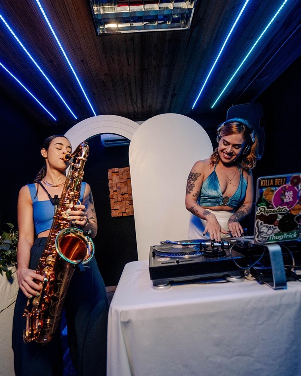 Foto do palco de uma festa em que é possível ver duas mulheres brancas, jovens, tocando instrumentos. A primeira toca um saxofone e a segunda mexe em picapes de DJs