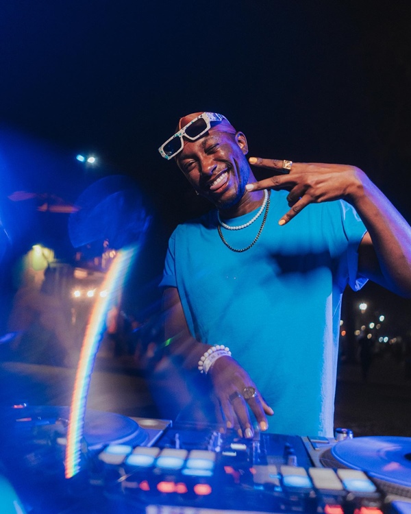 Homem jovem e negro, de cabelo raspado, tocando como DJ durante uma festa. Ele usa uma camiseta lisa e básica azul e um óculos escuro de armação branca