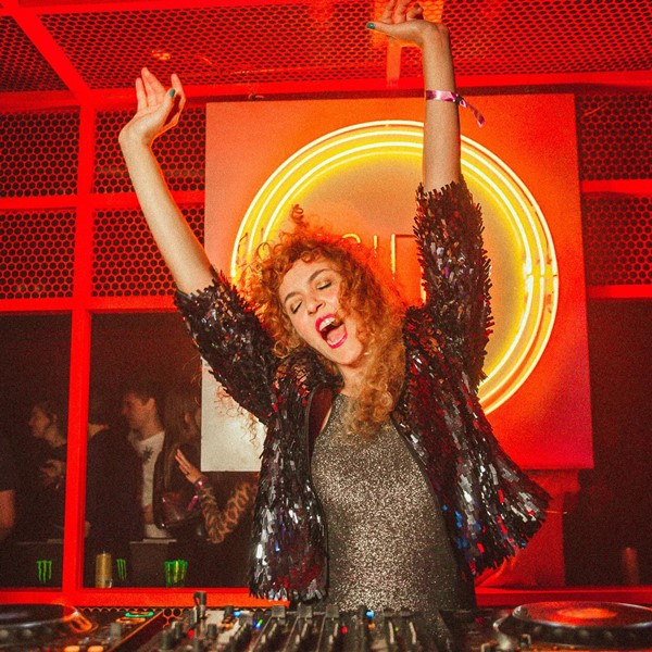 Mulher branca e ruiva, de cabelos longos cacheados, posando para foto em uma mesa de DJ durante uma festa