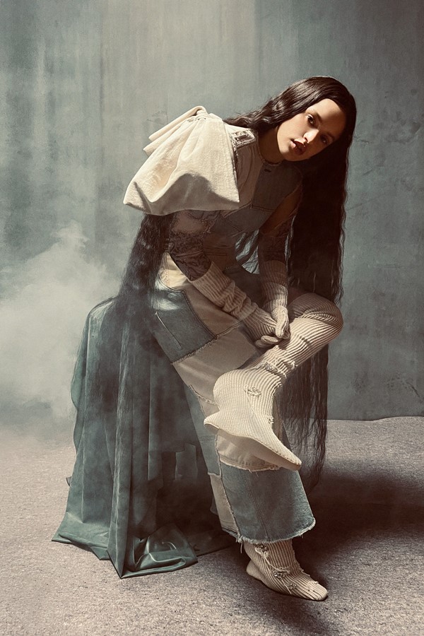 A cantora Rosalía, uma mulher jovem, branca, de cabelo ondulado longo, posando para a campanha da marca Acne Studios. Ela está em um estúdio com paredes marrons a e usa um conjunto de top e calça jeans, com retalhos do tecido em diferentes cores.