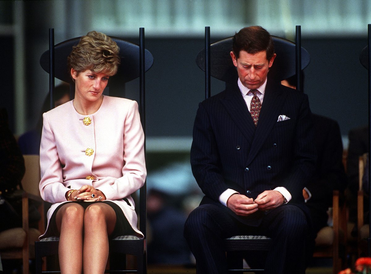 O príncipe Charles e a princesa de Gales, Lady Di, participam de uma cerimônia de boas-vindas em Toronto no início de sua turnê canadense, em outubro de 1991. Eles estão sentados em cadeiras pretas e olham para baixo. Ela usa um conjunto rosa de blazer e saia e ele terno preto com camisa branca.