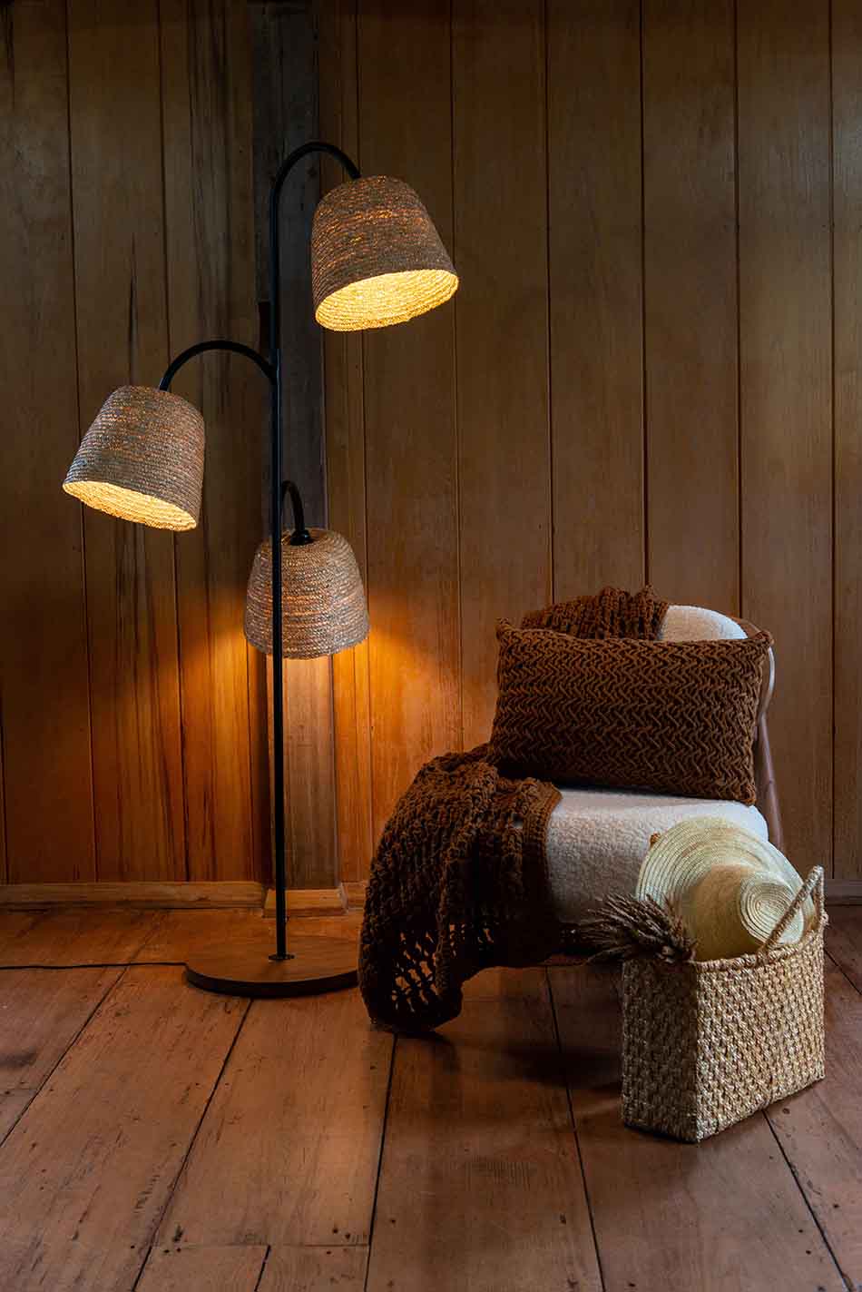 Foto da decoração de uma casa de madeira. Na imagem é possível ver uma luminária de ferro preta, uma poltrona branca com uma manta de crochê marrom por cima e, ao lado, uma bolsa estilo cesto de palha.