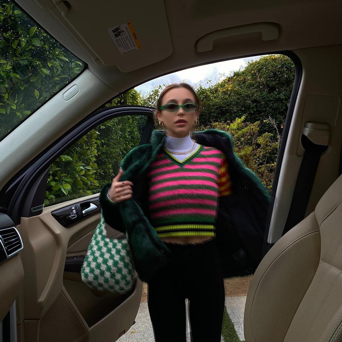 A influencer Emma Chamberlain, uma mulher branca e jovem, com cabelo loiro liso, posando para foto na porta de um carro preto. Usa uma regata de gola alta branca, um colete listrado colorido, um casaco verde escuro e um óculos escuro verde.