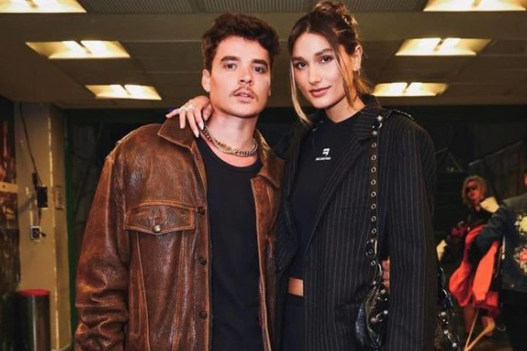 João Figueiredo de camiseta preta e jaqueta marrom posa abraçado em Sasha, que veste saia e blusa preta e blazer preto com uma bolsa pendurada, um coque e fios soltos na frente do rosto no estilo
