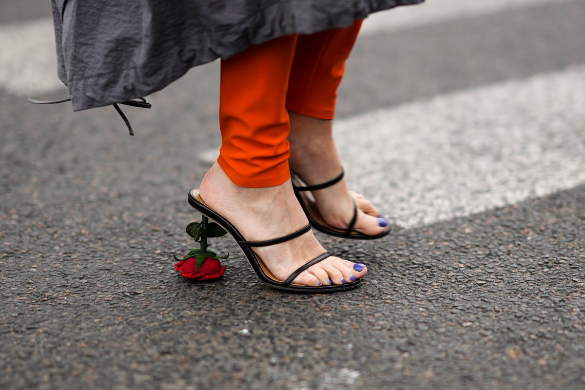 Mulher branca andando pelas ruas de Paris, na França. Na foto é possível ver apenas seus pés, que calçam uma sandália de salto da marca Loewe. O item é preto e de couro e possui um salto em formato da flor rosa. Usa ainda uma calça laranja e um casaco cinza.