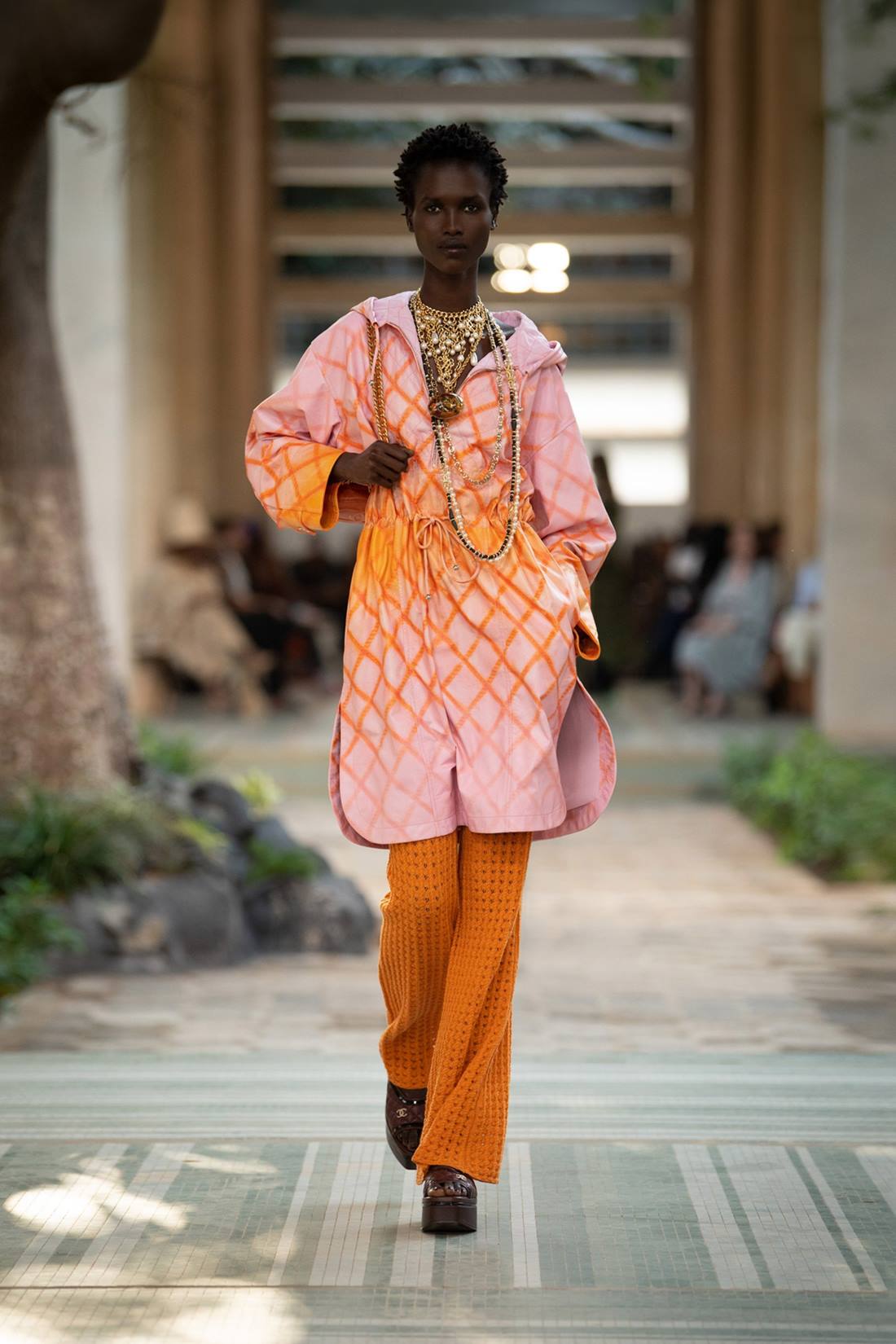 Modelo negra e jovem, com cabelo curto cacheado, andando na passarela do desfile Pre-Fall 2023 da marca Chanel. Ela usa uma calça laranja folgada e uma camisa estilo bata rosa com listras laranjas. - Metrópoles