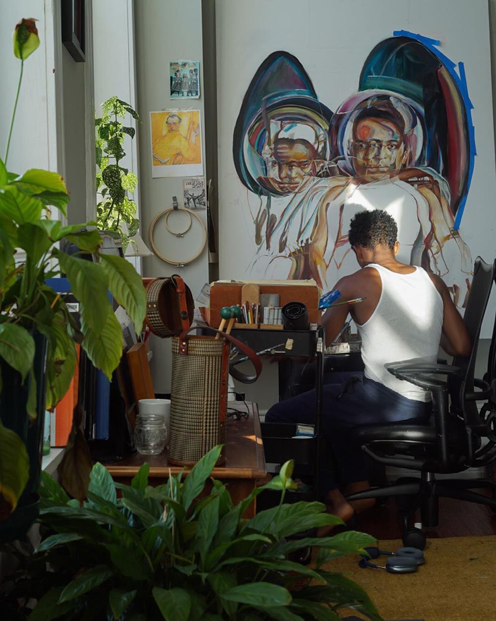 O artista plástico Chris Pleasant, um homem negro e jovem, de cabelo curto, em seu estúdio de pintura. Usa uma camiseta regata branca e calças pretas e está sentado em uma cadeira enquanto desenha. - Metrópoles