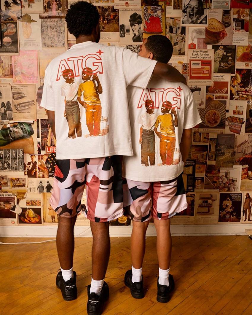 Dois homens jovens e negros posam para foto em frente a uma parede cheia de colagens. A imagem é uma campanha publicitária da marca All Things Good. Eles usam uma camiseta branca com uma pintura atrás e uma bermuda estampada. - Metrópoles