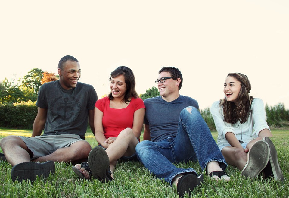 Grupo de quatro pessoas jovens rindo sentadas em grama - Metrópoles