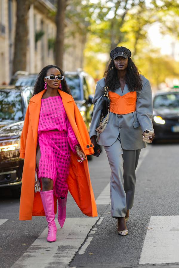 Street style: em faixa de pedestre, amigas caminham lado a lado. Uma delas usa look rosa com sobretudo laranja. A outra veste conjunto cinza de alfaiataria com sobreposição de corset laranja - Metrópoles
