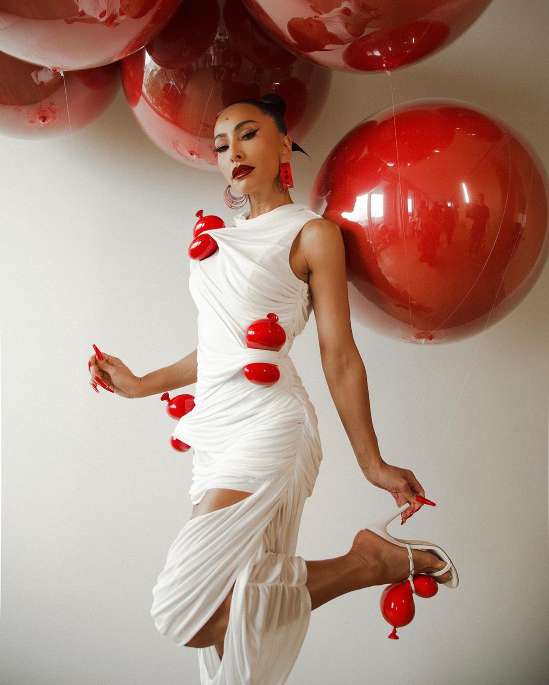 A ex-BBB e apresentadora Sabrina Sato, uma mulher jovem e morena, com traços asiáticos, posa para foto em um fundo branco com balões vermelhos. Usa um vestido branco com balões vermelos amarrados e uma sandália branca, de tiras, também com um balão vermelho amarrado. - Metrópoles